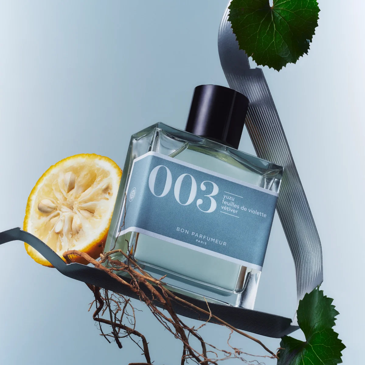 Bon Parfumeur - Les Classiques - 003 Flakon mit blauem Etikett, Zitrusfrucht und grünen Blättern vor hellblauem Hintergrund