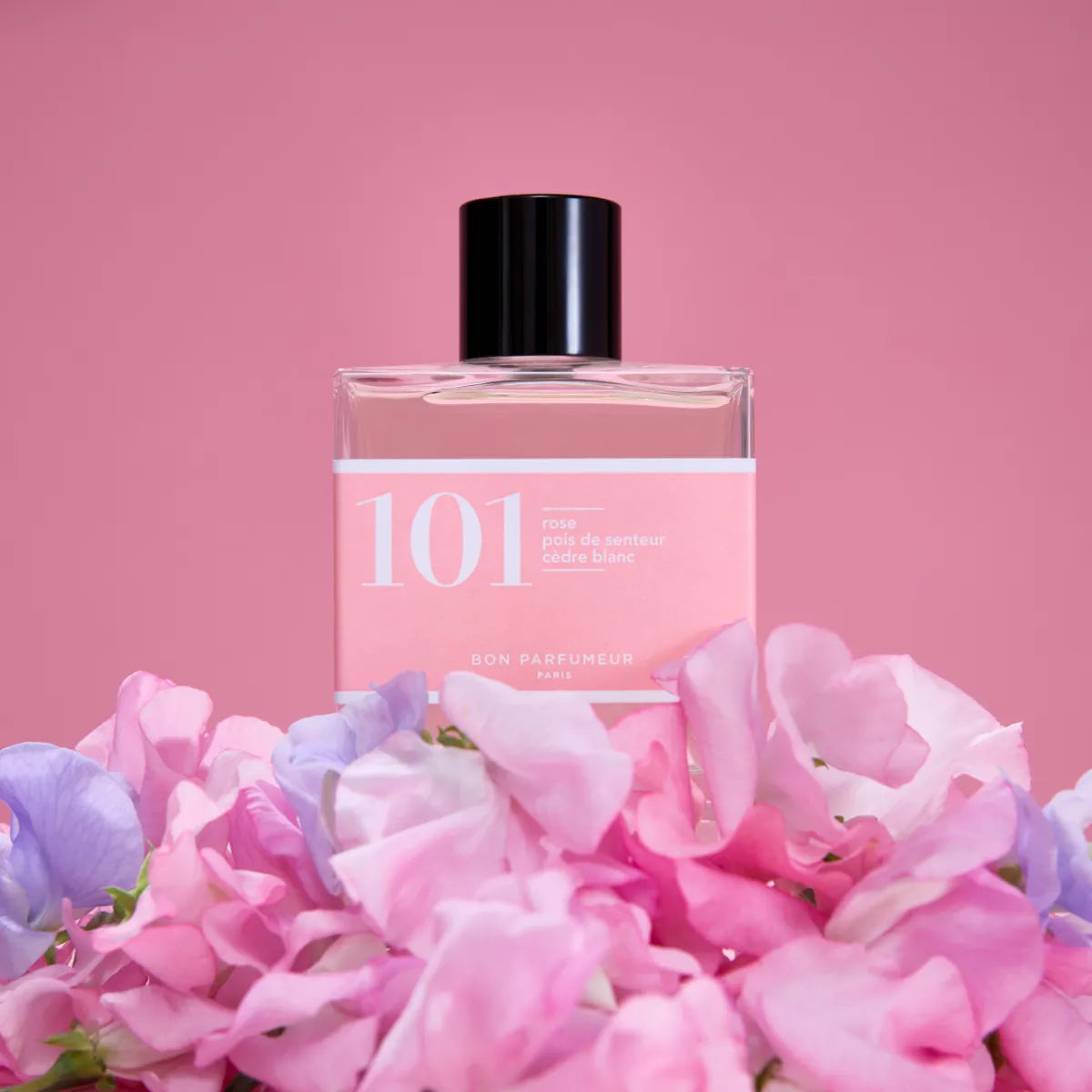 Bon Parfumeur - Les Classiques - 101 Flakon mit rosa Etikett und schwarzem Verschluss umgeben von rosa und lila Blüten vor rosafarbenem Hintergrund