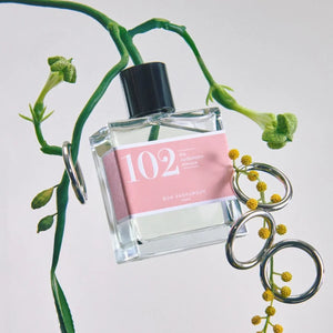 Bon Parfumeur - Les Classiques - 102 Parfümflakon mit rosa Etikett und schwarzem Verschluss zwischen grünen Zweigen und kleinen gelben Blüten