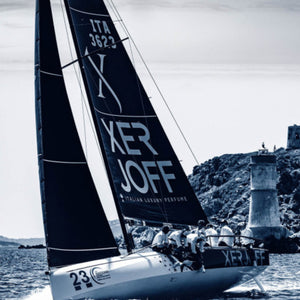 Xerjoff - Join The Club - 40 Knots Segelboot mit schwarzem Segel und Bootscrew vor mediterraner Küste bei sonnigem Wetter