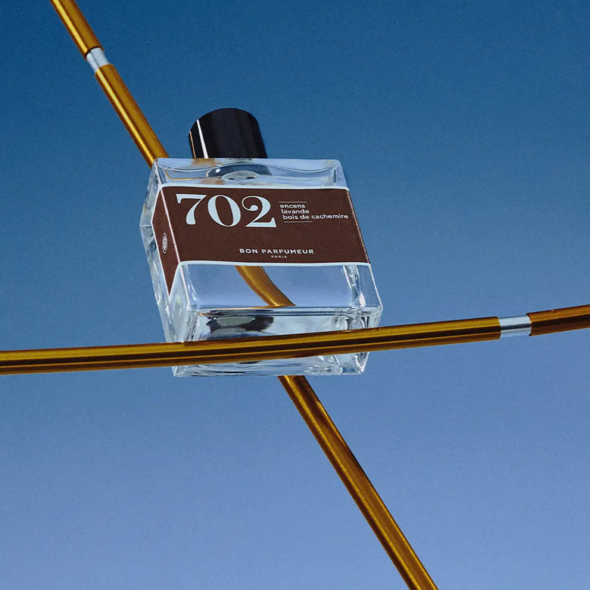 Bon Parfumeur - Les Classiques - 702 Parfumflakon mit klarem Glas und braunem Etikett vor blauem Himmel und goldenen Stäben