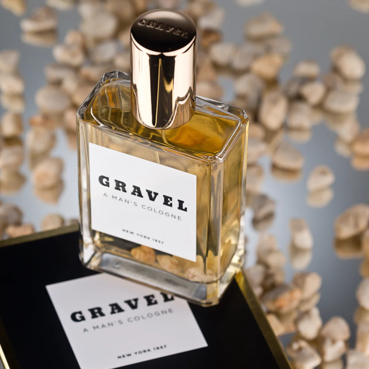 Gravel - A Man's Cologne Parfumflakon mit goldfarbenem Verschluss und Kieselsteinen in klarem Glas vor spiegelnder Oberfläche