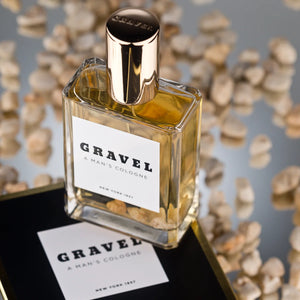 Gravel - A Man's Cologne Parfumflakon mit goldfarbenem Verschluss und Kieselsteinen in klarem Glas vor spiegelnder Oberfläche