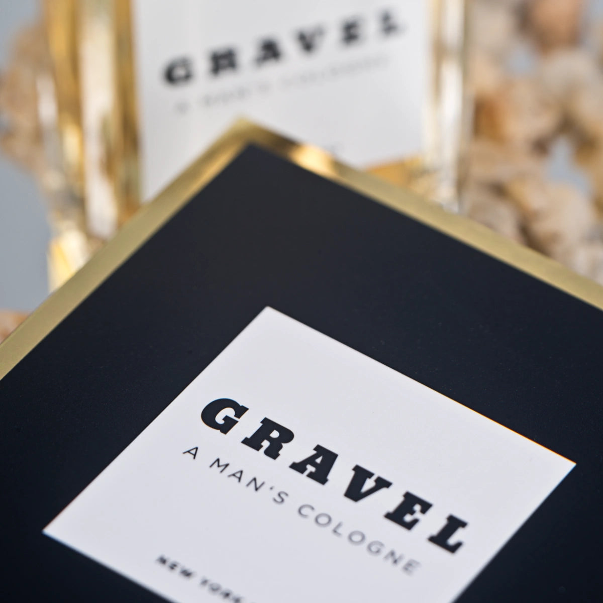 Gravel - A Man's Cologne schwarze Verpackung mit weißem Etikett und goldfarbenem Rand vor unscharfem Hintergrund