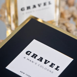Gravel - A Man's Cologne schwarze Verpackung mit weißem Etikett und goldfarbenem Rand vor unscharfem Hintergrund