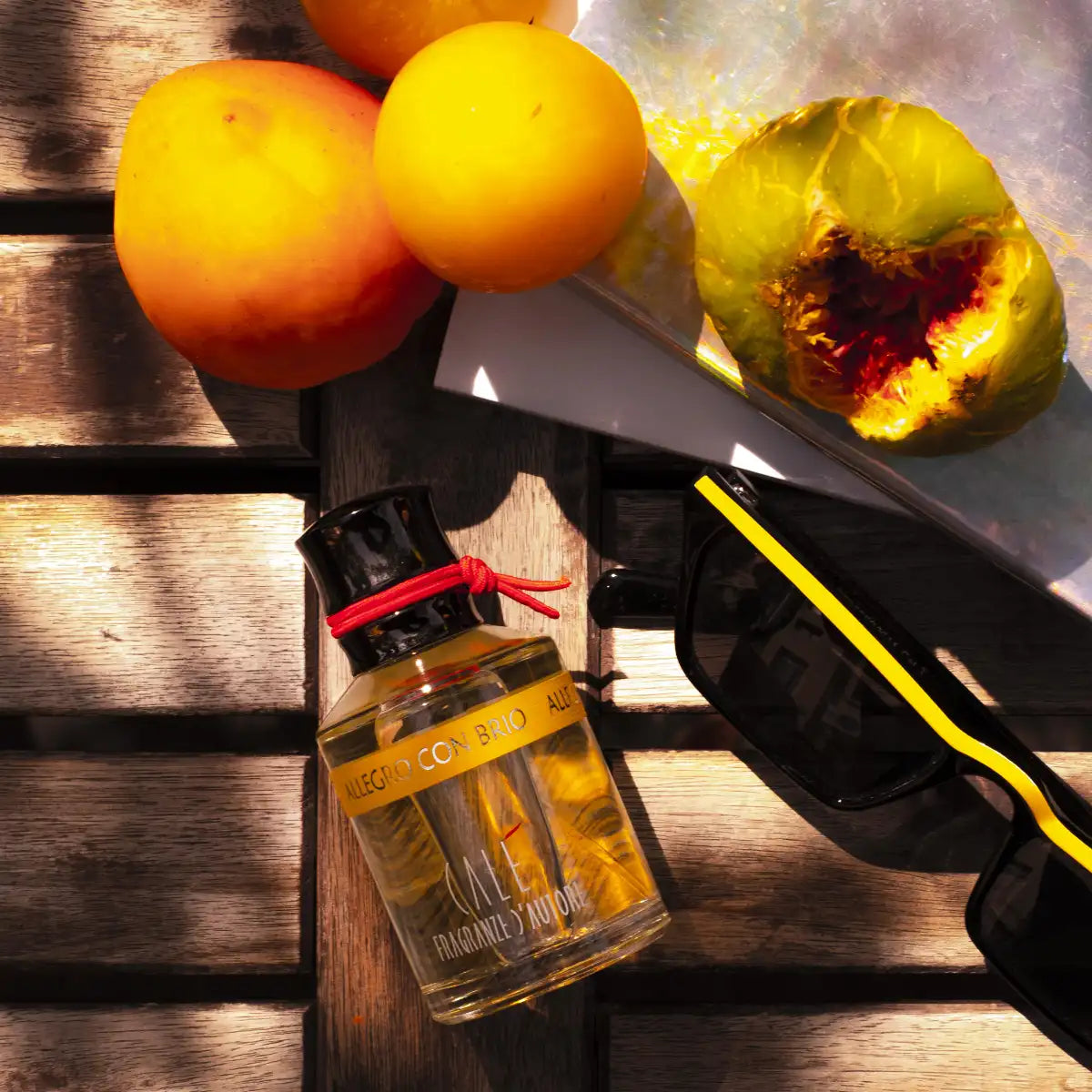 Calé Fragranze d'Autore - Allegro con Brio Flakon mit gelbem Etikett neben Sonnenbrille und Obst auf Holzbohlen