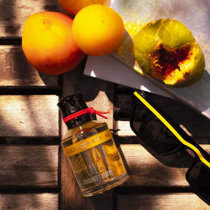 Calé Fragranze d'Autore - Allegro con Brio Flakon mit gelbem Etikett neben Sonnenbrille und Obst auf Holzbohlen