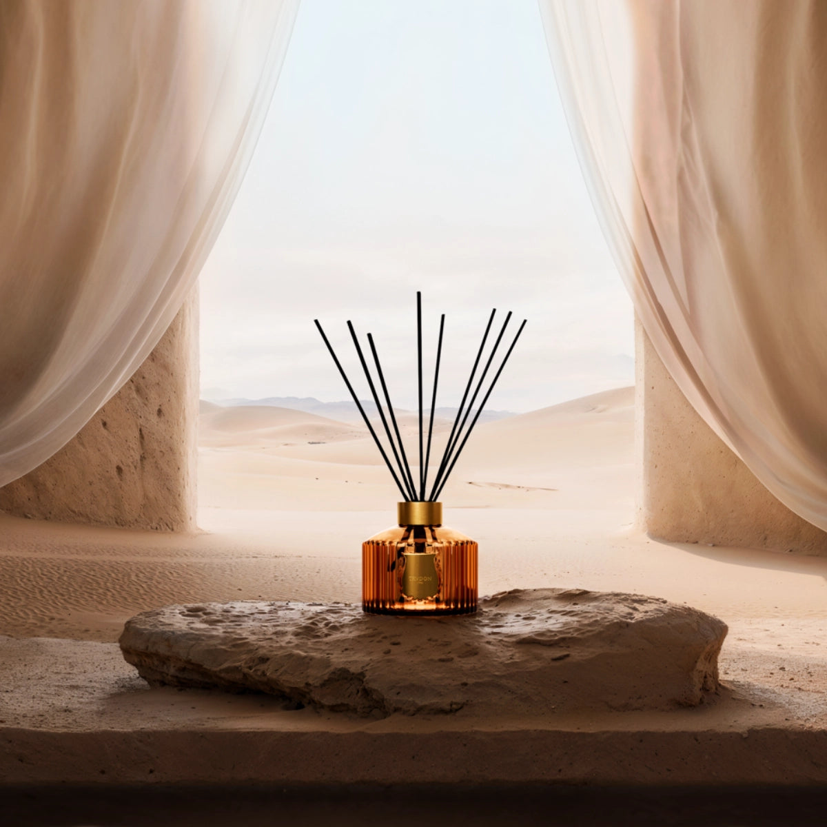 Trudon - Altaïr - Diffusor mit bernsteinfarbener Flasche und schwarzen Stäben vor Wüstenlandschaft und hellem Vorhang