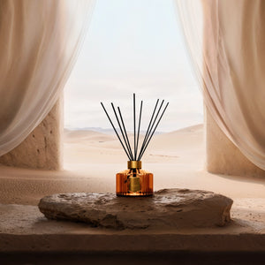 Trudon - Altaïr - Diffusor mit bernsteinfarbener Flasche und schwarzen Stäben vor Wüstenlandschaft und hellem Vorhang