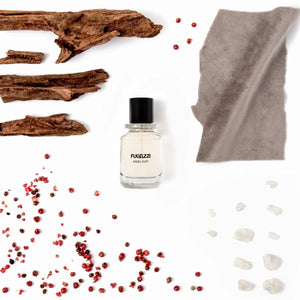 Fugazzi - Angel Dust Extrait Parfümflakon mit schwarzen Deckel umgeben von Holzstücken und rosa Pfefferkörnern auf weißem Grund