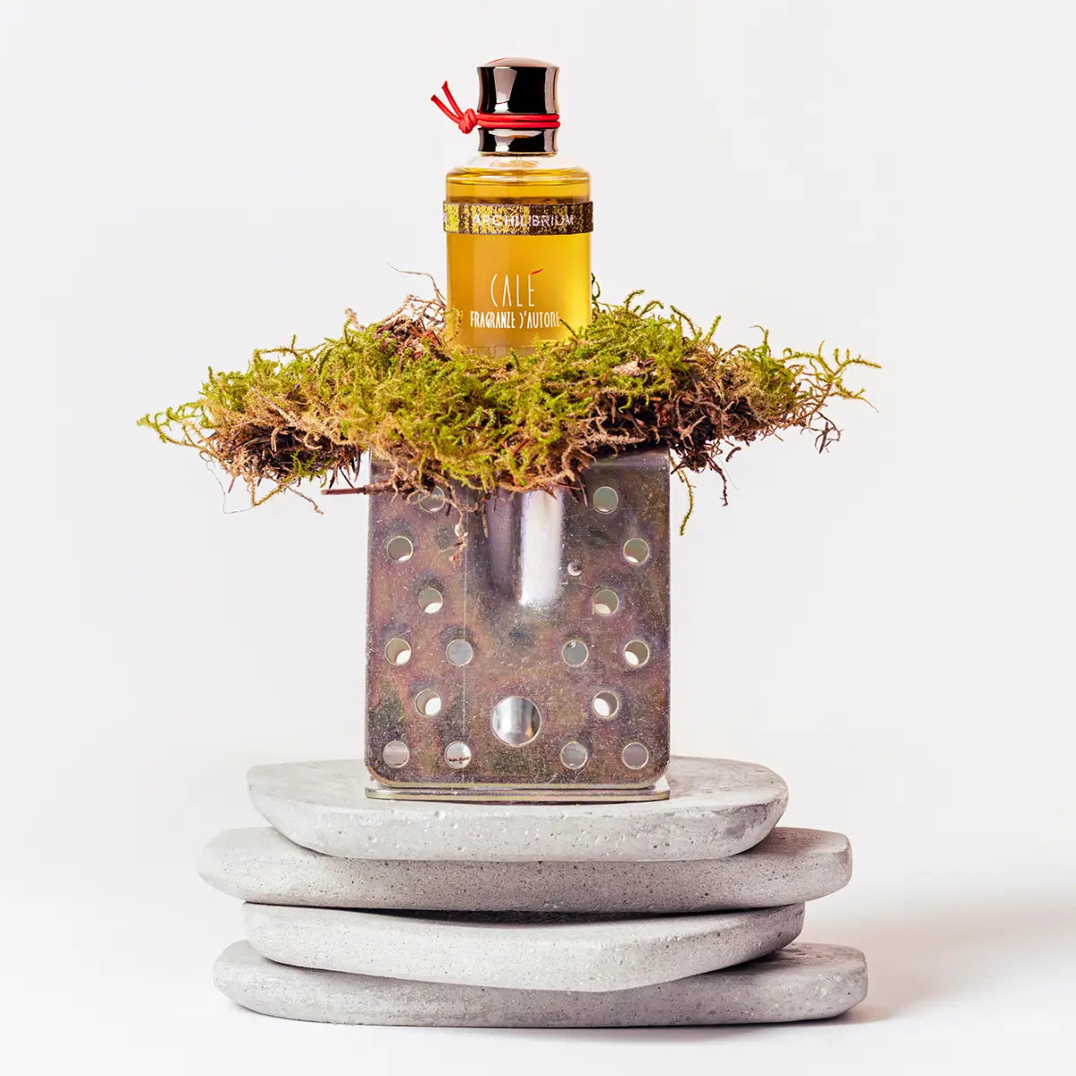 Calé Fragranze d'Autore - Archilibrium Flakon mit goldenem Inhalt auf Moos und geometrischen Steinen vor weißem Hintergrund