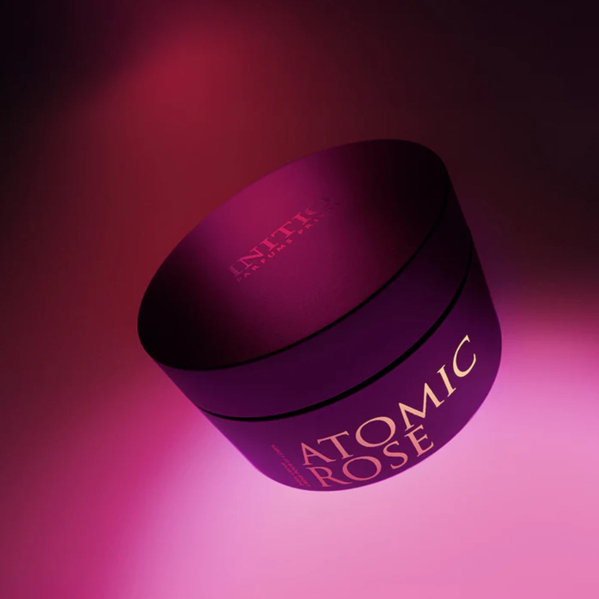 Initio - Atomic Rose - Body Cream elegante dunkelviolette Dose mit goldener Schrift vor pinkem Lichtschein