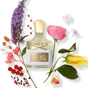Creed - Aventus for Her Flakon mit goldenem Verschluss und floralen Elementen vor hellem Hintergrund