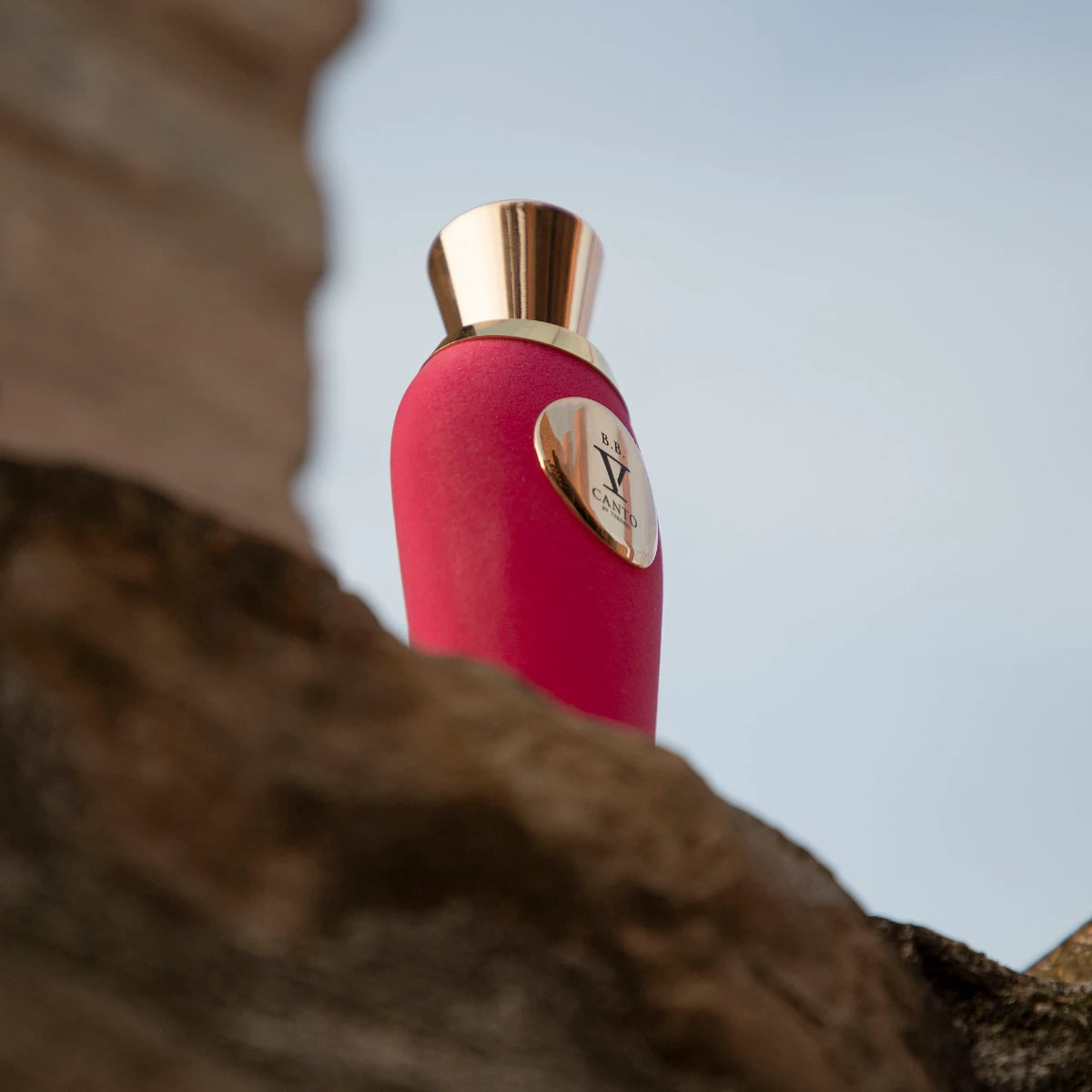 V Canto - B.B. zeigt eleganten pinken Flakon mit goldfarbenem Verschluss und rundem Logo vor hellem Himmel