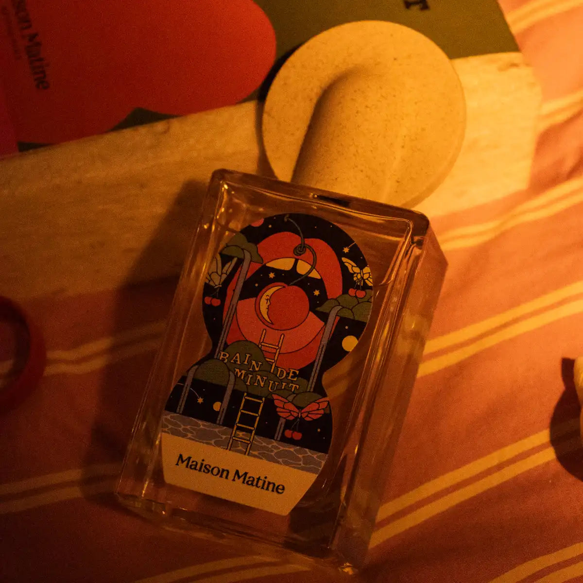 Maison Matine - Night Fever Collection - Bain de Minuit zeigt Flakon mit farbiger Illustration in warmem Licht auf Stoffoberfläche