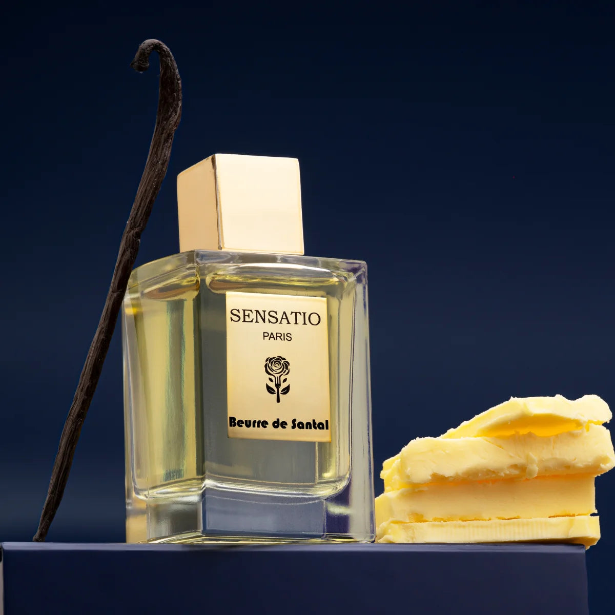 Sensatio Paris - Beurre de Santal Parfumflakon mit goldfarbenem Verschluss, Vanilleschote und Butterstücken vor dunklem Hintergrund