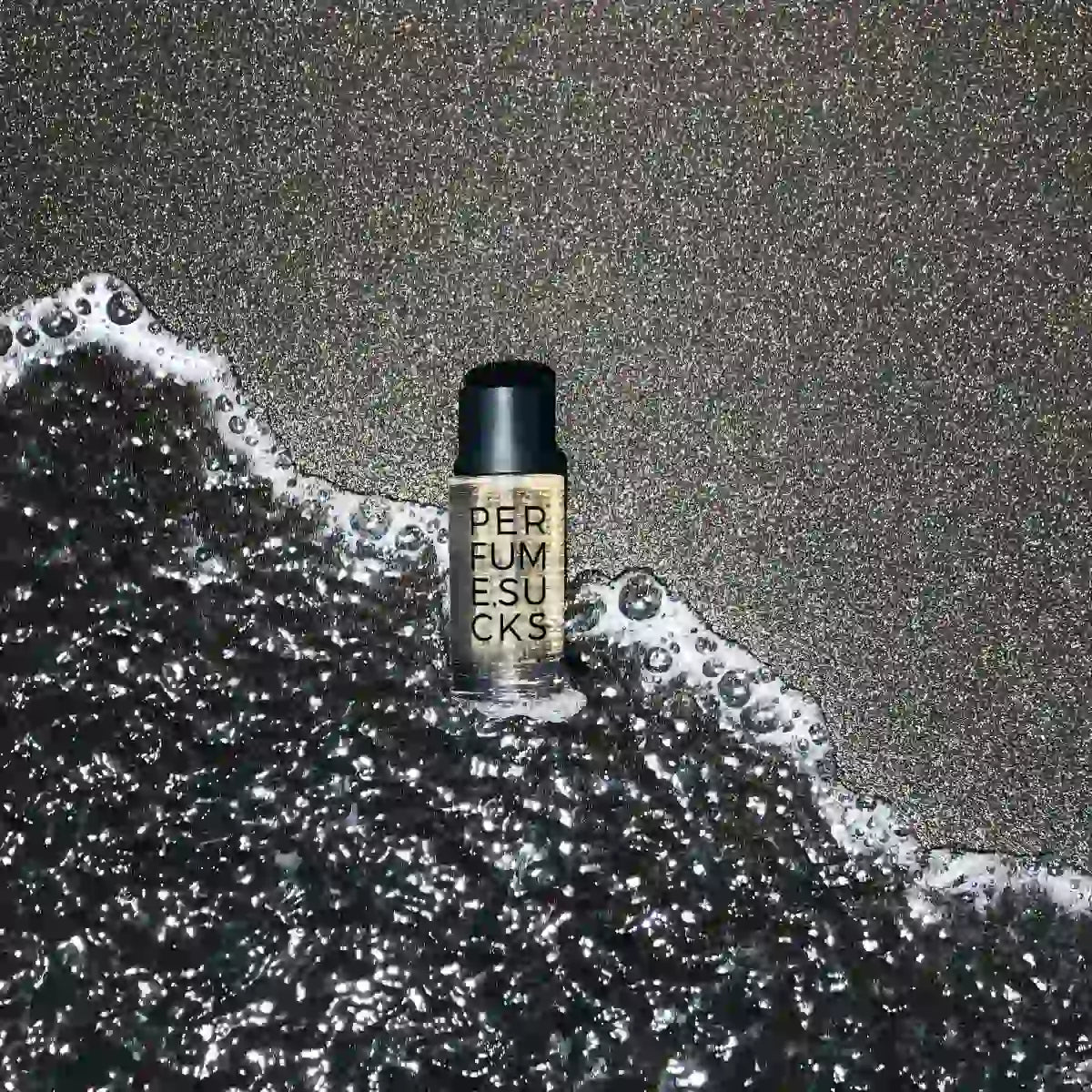 PERFUME.SUCKS - Black C Flakon mit schwarzem Verschluss und klarer Schrift auf schwarzem Sand mit Meereswellen