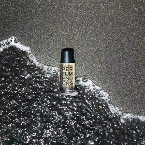 PERFUME.SUCKS - Black C Flakon mit schwarzem Verschluss und klarer Schrift auf schwarzem Sand mit Meereswellen