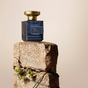 Atelier Materi - Black Oregano zeigt dunkelblauen Flakon mit goldenem Verschluss auf grobem Stein und getrockneten Kräutern