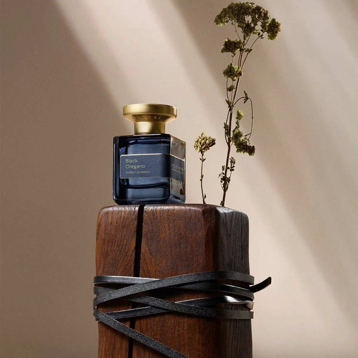 Atelier Materi - Black Oregano zeigt dunkelblauen Flakon mit goldenem Verschluss auf Holzsockel und getrocknetem Zweig