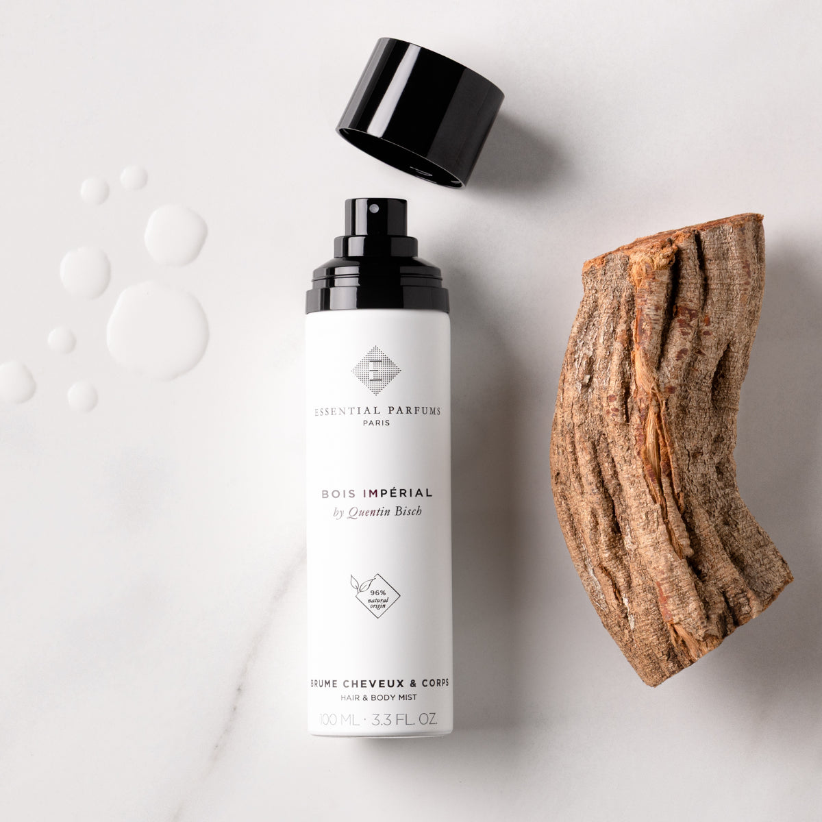 Essential Parfums - Bois Impérial - Hair & Body Mist weiße Flasche mit offenem schwarzen Deckel neben Holzstück auf hellem Hintergrund