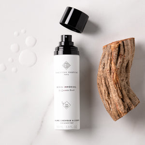 Essential Parfums - Bois Impérial - Hair & Body Mist weiße Flasche mit offenem schwarzen Deckel neben Holzstück auf hellem Hintergrund