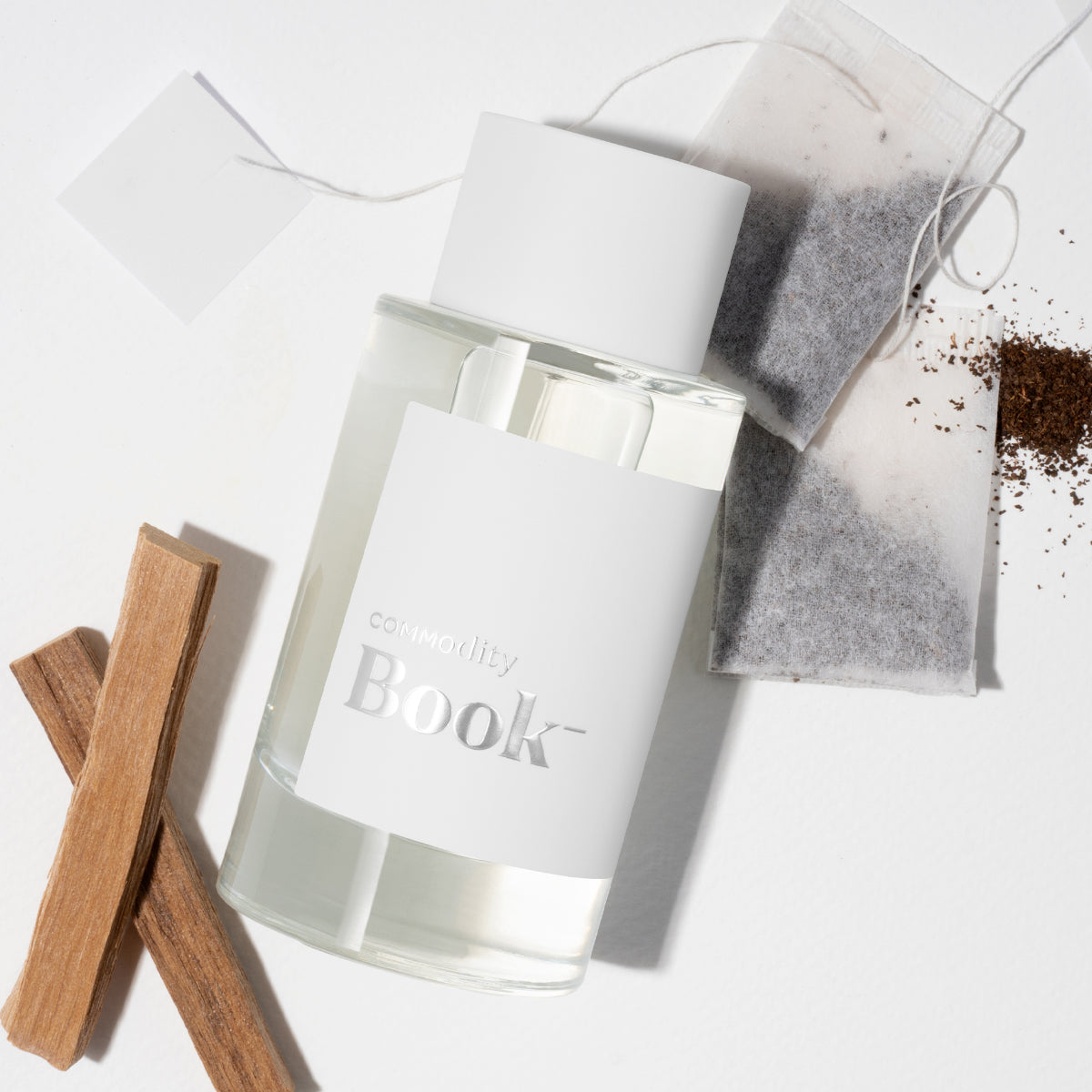 Commodity - Book- Parfumflakon mit weißem Etikett, Tee-Beuteln und Holzstücken auf hellem Hintergrund inszeniert