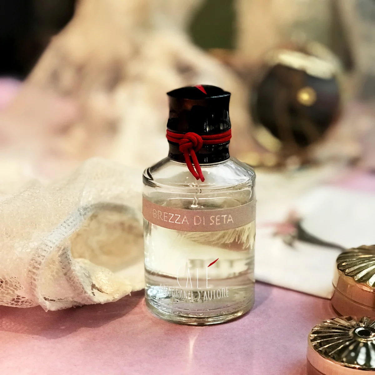 Calé Fragranze d'Autore - Brezza di Seta Glasflakon mit rotem Band auf rosa Oberfläche neben zartem Stoff