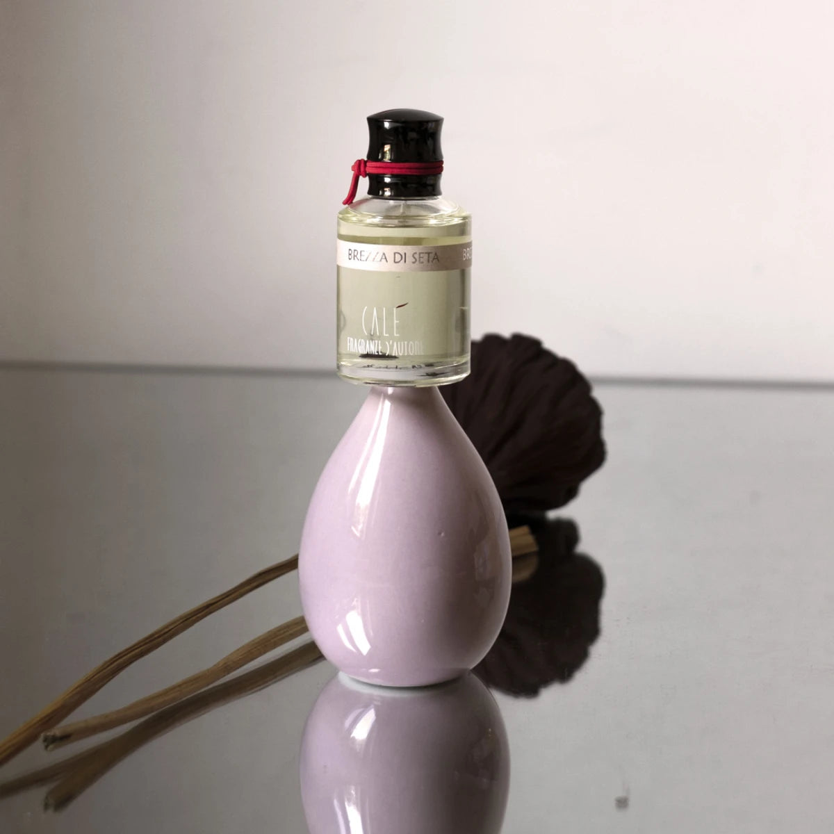 Calé Fragranze d'Autore - Brezza di Seta Flakon auf zartrosa Keramikvase mit dunklem dekorativem Element im Hintergrund