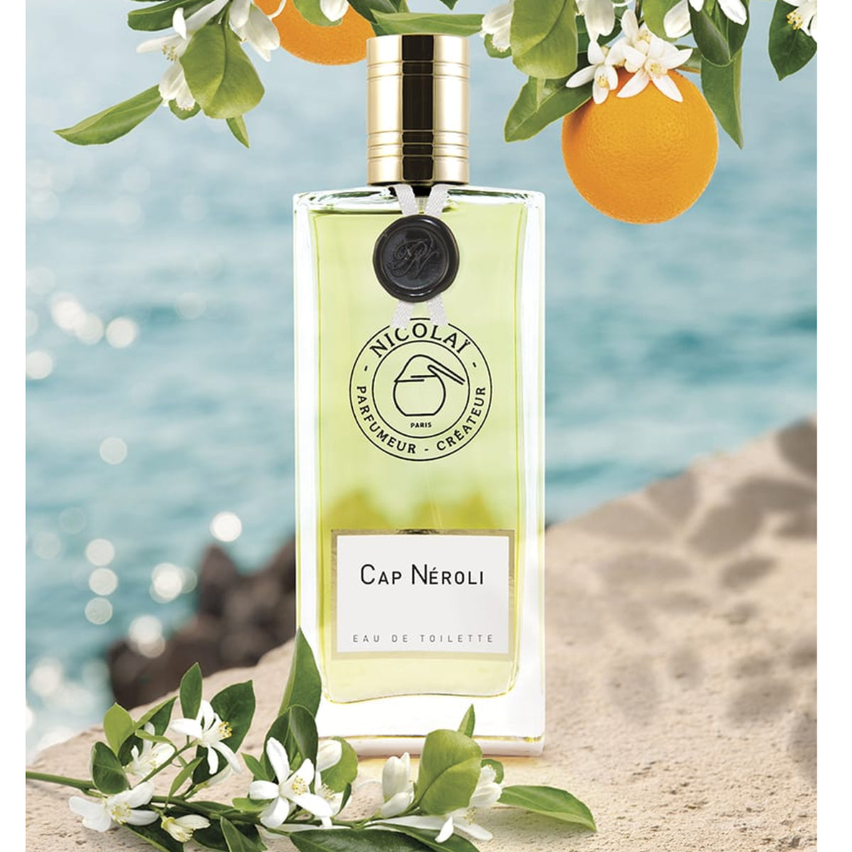 Nicolaï - Cap Néroli Eau de Toilette mit goldenem Verschluss und grüner Flüssigkeit vor Meereskulisse und Orangenblüten