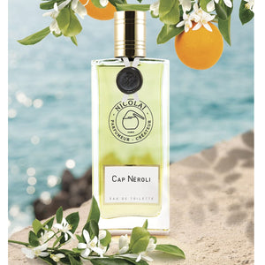 Nicolaï - Cap Néroli Eau de Toilette mit goldenem Verschluss und grüner Flüssigkeit vor Meereskulisse und Orangenblüten