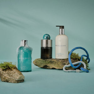 Molton Brown - Coastal Cypress & Sea Fennel Hand Lotion weiße Flasche mit schwarzem Pumpspender vor grün-blauem Hintergrund mit Felsen und Kletterseil