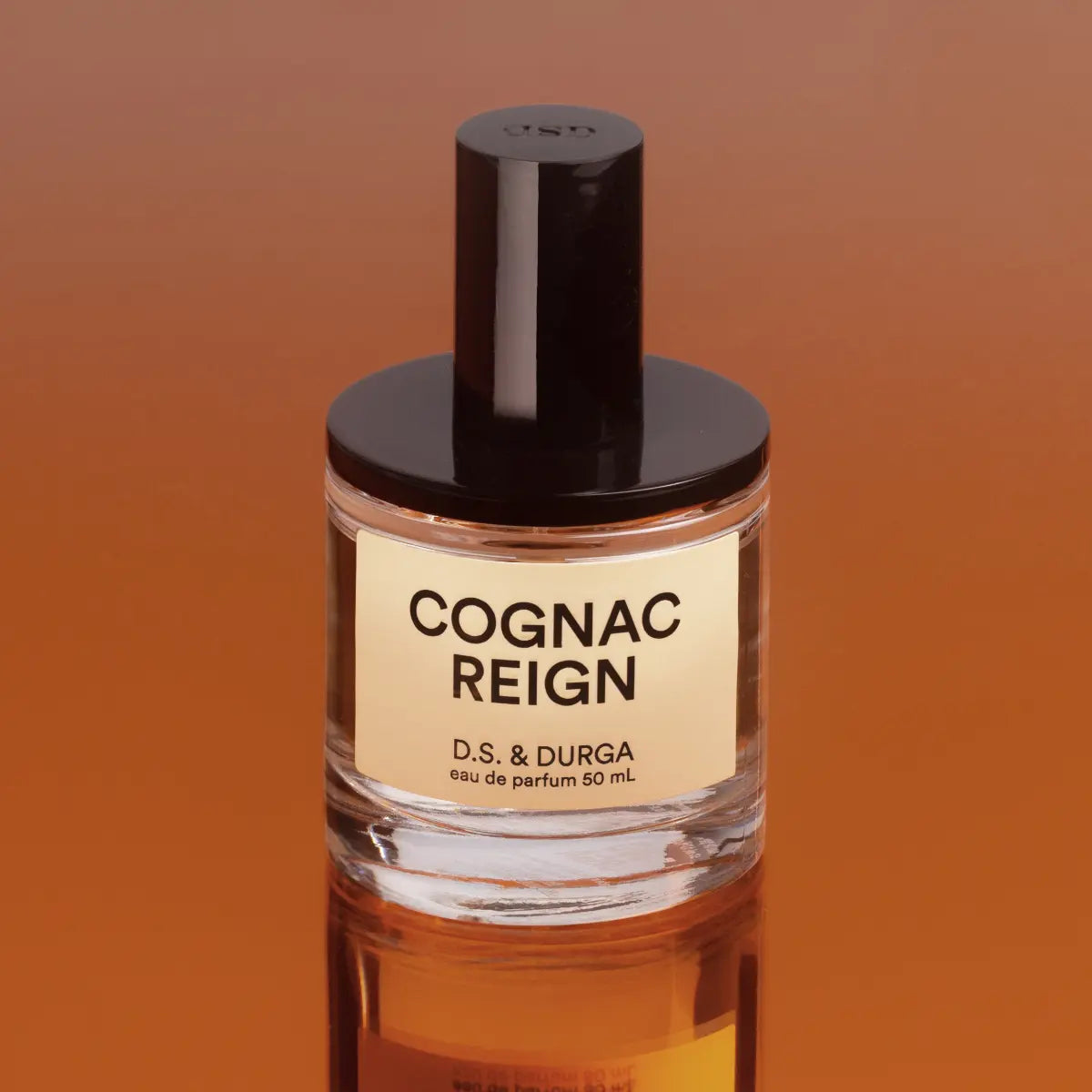 D.S. & Durga - Cognac Reign Parfumflakon mit schwarzem Verschluss vor reflektierendem cognacfarbenem Hintergrund