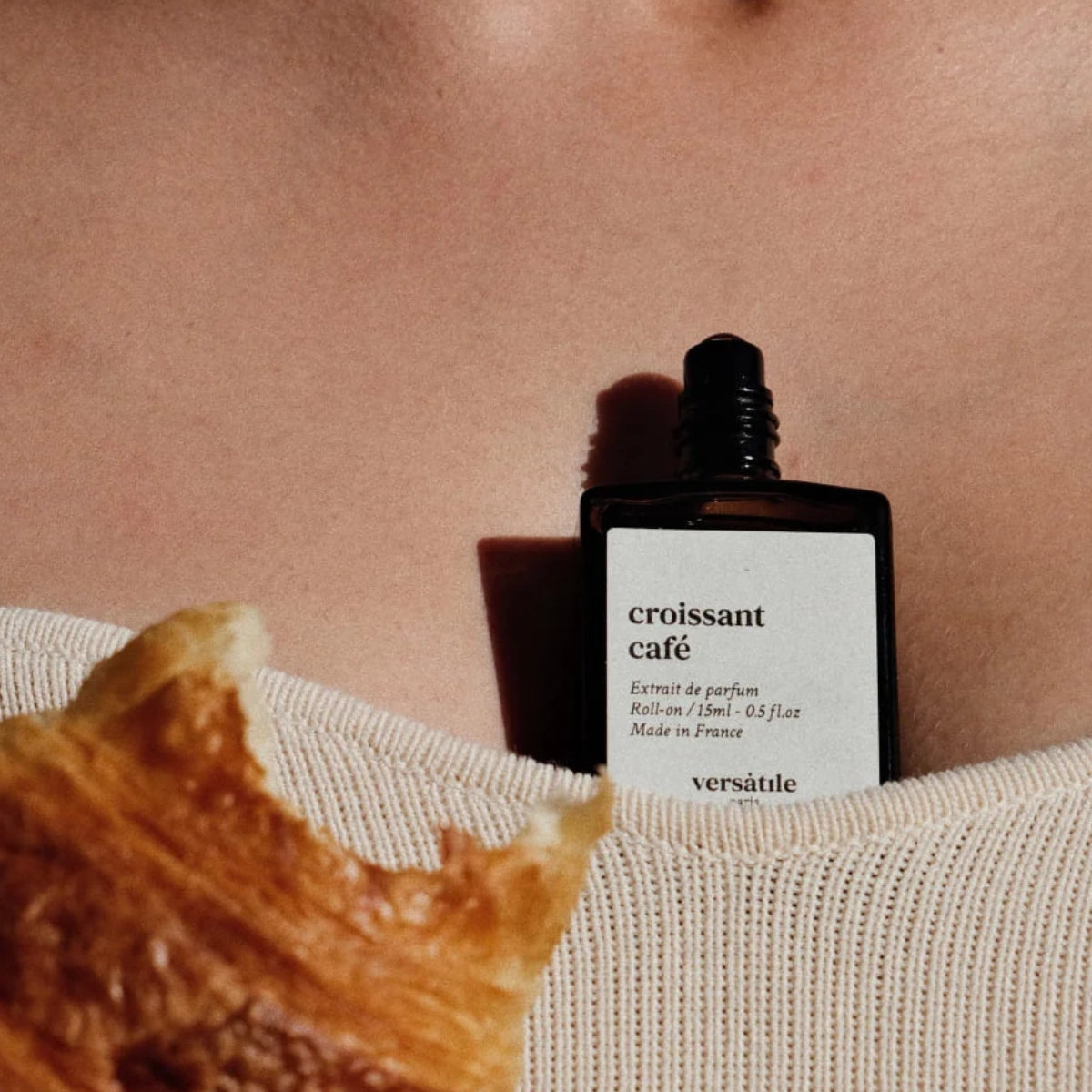 Versatile - Croissant Café Roll-on Flakon auf Haut stehend neben einem goldbraunen Croissant in natürlichem Licht