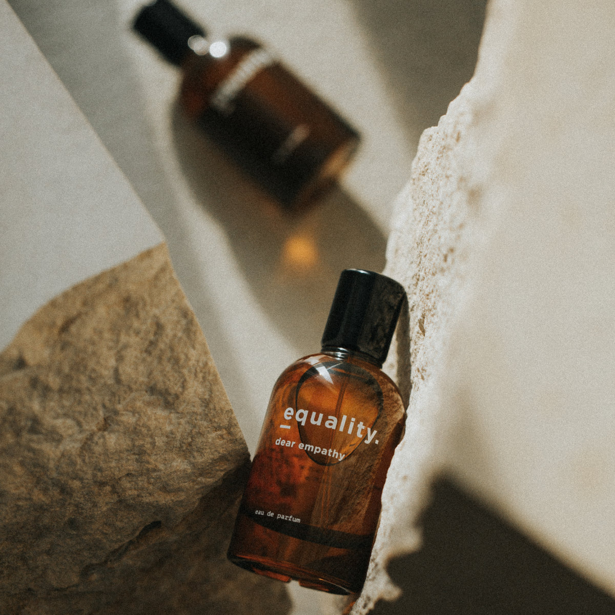 equality. fragrances - dear empathy Parfumflakon in bernsteinfarbenem Glas mit schwarzem Verschluss auf hellem Steinuntergrund