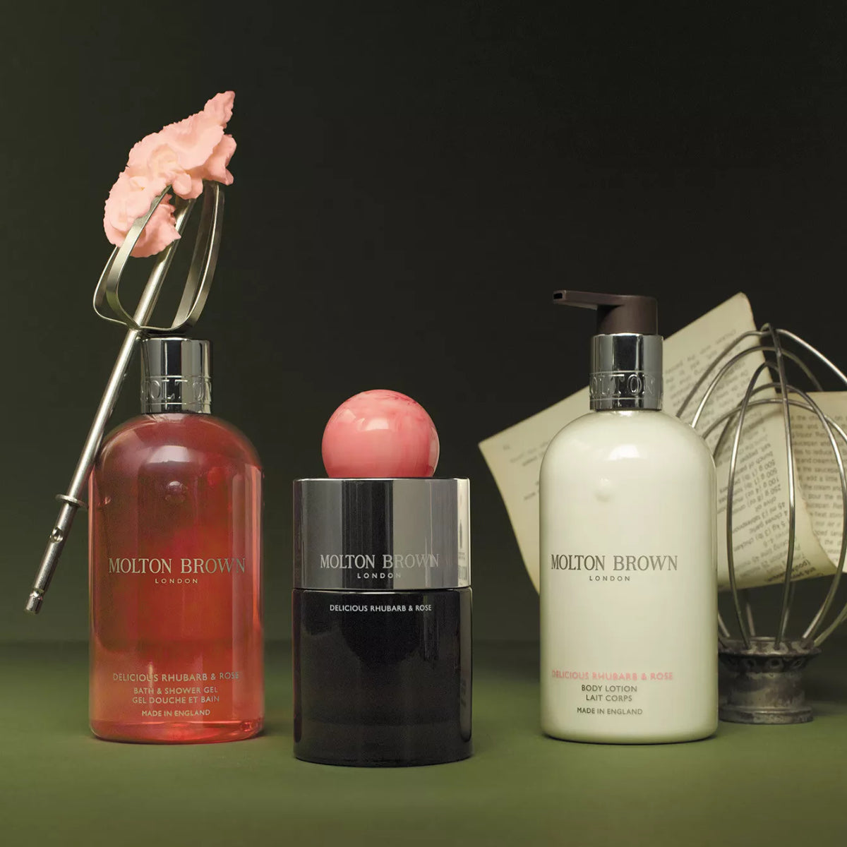 Molton Brown - Delicious Rhubarb & Rose Hand Lotion im cremefarbenen Spender vor Küchenutensilien und grünem Hintergrund