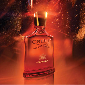 Creed - Delphinus Parfümflakon in leuchtendem Orange mit goldenem Verschluss und funkelndem Amber-Effekt im Hintergrund