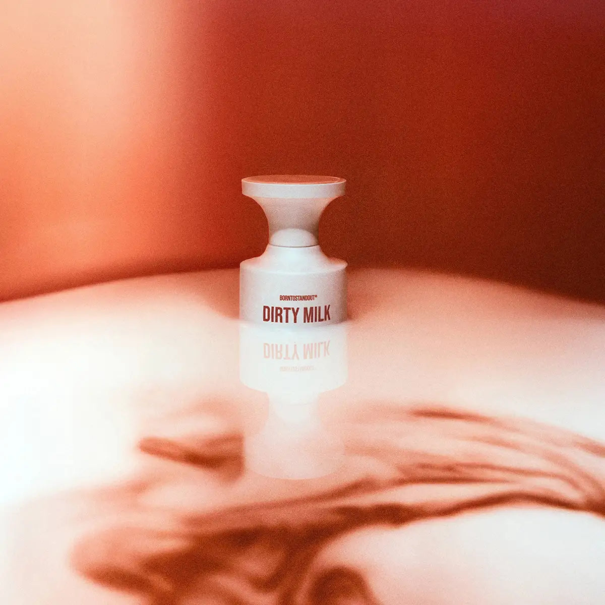 BORNTOSTANDOUT - Dirty Milk zeigt weißen Flakon mit roter Schrift auf glänzendem, warmem Untergrund mit Spiegelung.