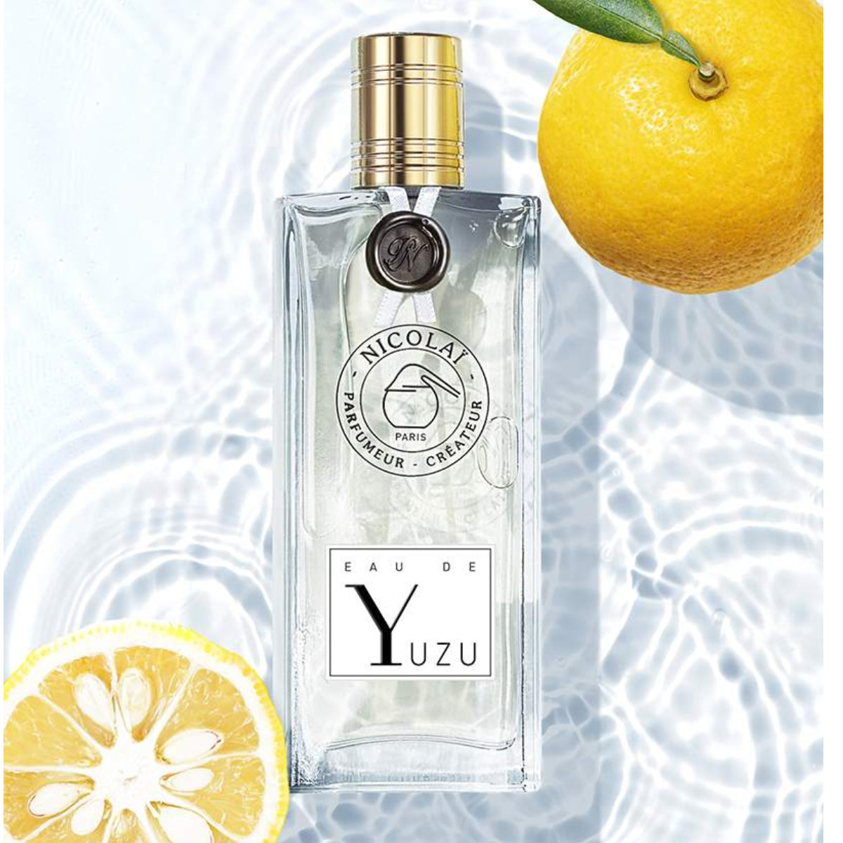 Nicolaï - Eau de Yuzu Parfumflakon mit klarem Glas und goldfarbenem Verschluss neben Yuzu-Früchten im Wasserlicht