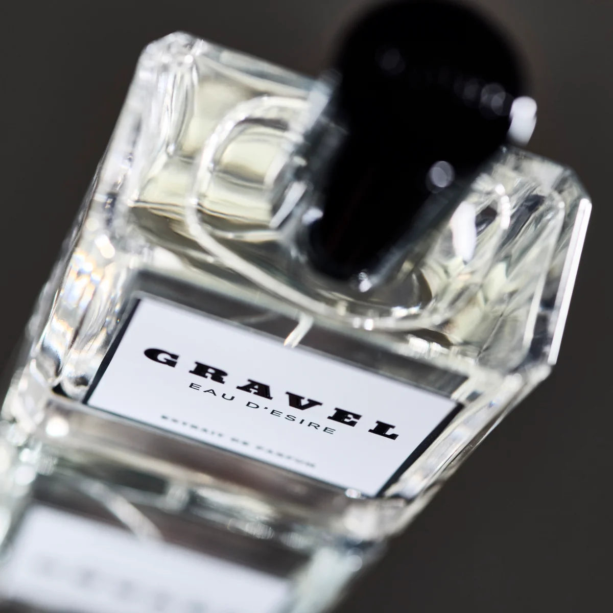 Gravel - Eau D'Esire Parfumflakon mit klarem Glas und schwarzem Verschluss auf dunklem, unscharfem Hintergrund