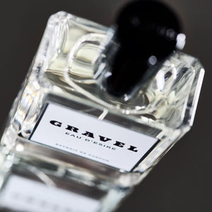 Gravel - Eau D'Esire Parfumflakon mit klarem Glas und schwarzem Verschluss auf dunklem, unscharfem Hintergrund