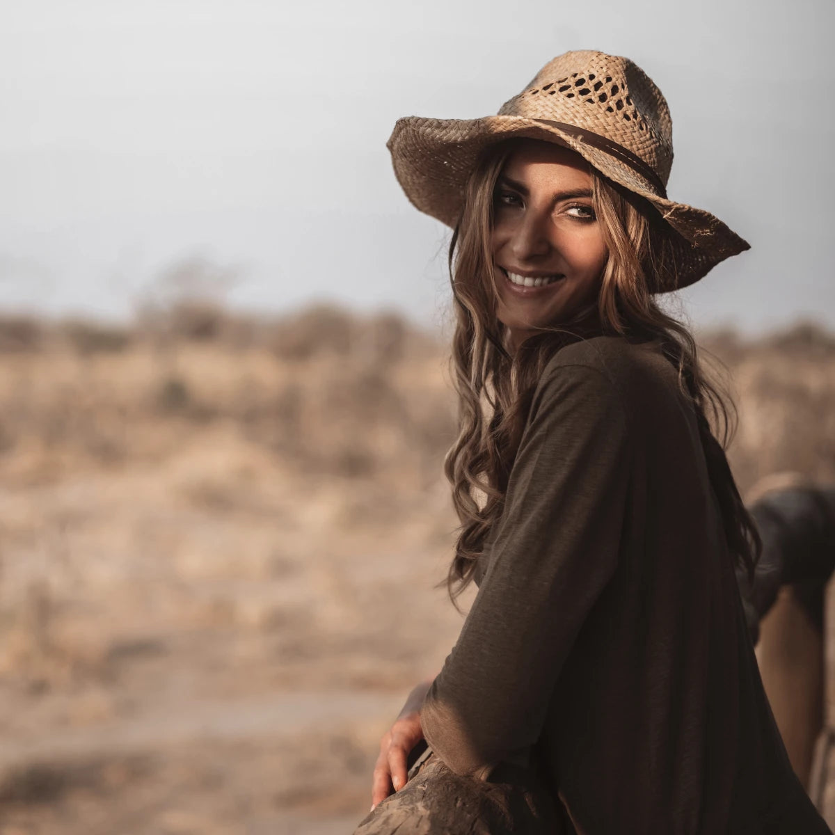 Frau mit Strohhut und lockerem braunem Oberteil lächelnd in natürlicher, sonnendurchfluteter Landschaft Ella K - Ghibli
