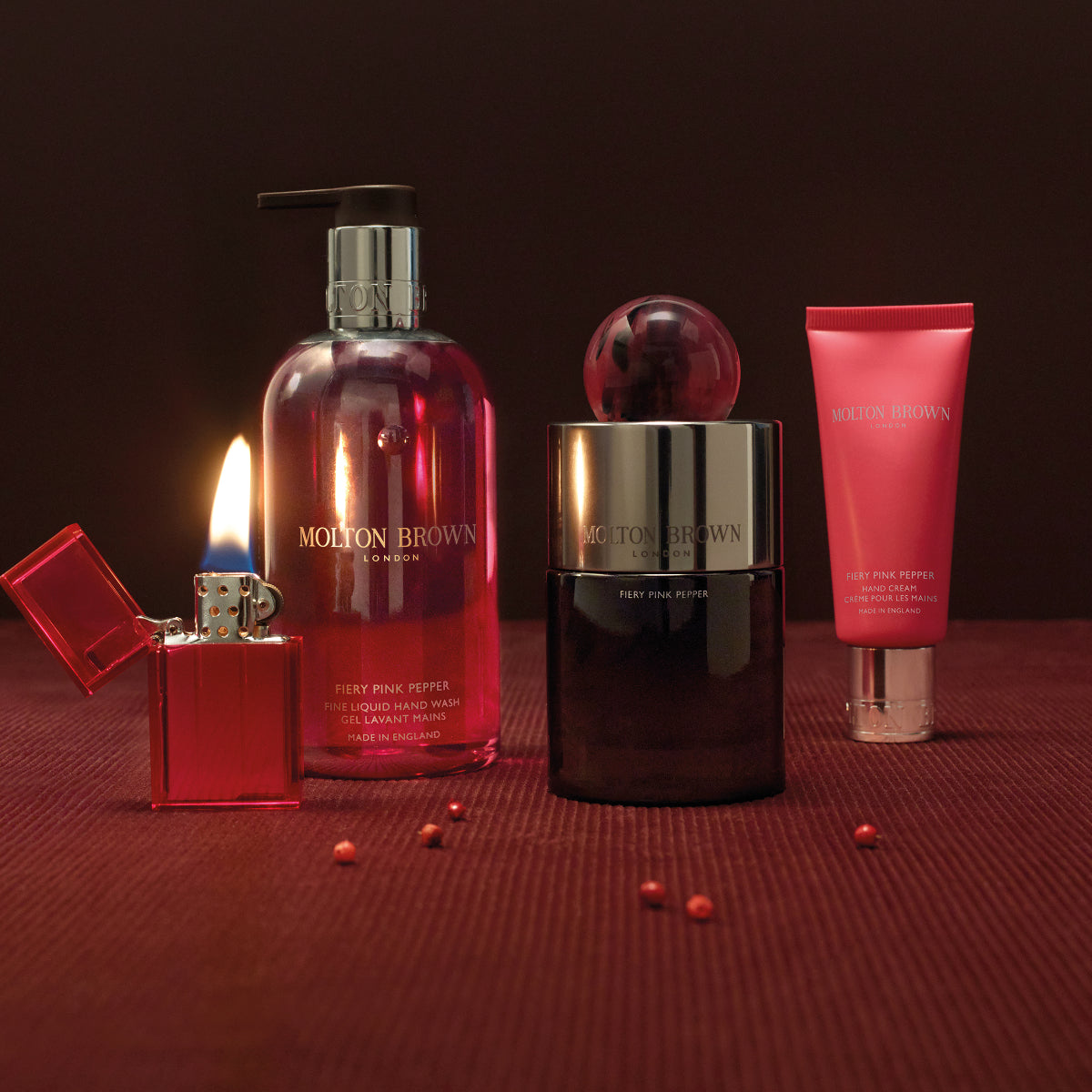 Molton Brown - Fiery Pink Pepper - Hand Cream in roter Tube neben Parfumflakon und Handseife vor dunklem Hintergrund