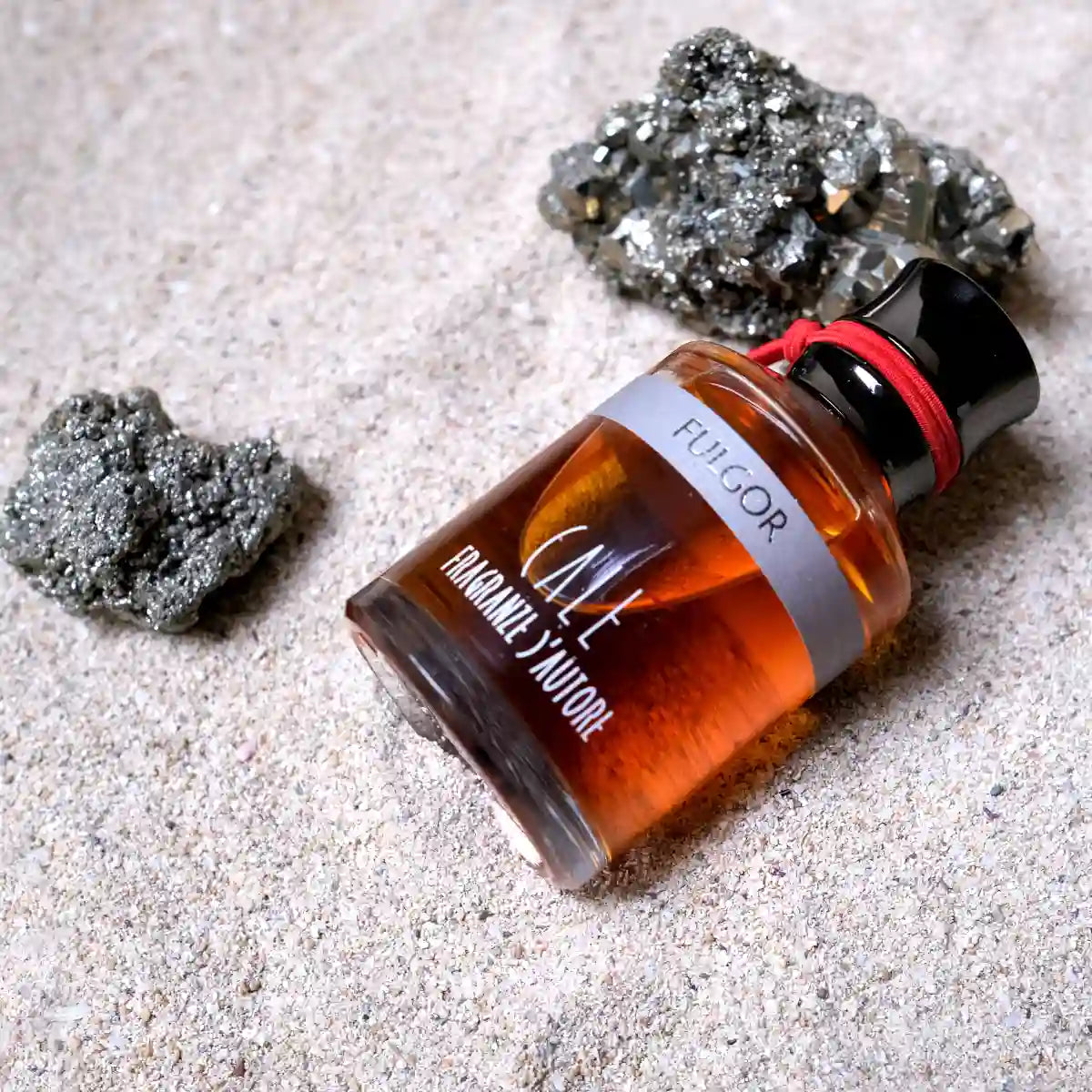 Calé Fragranze d'Autore - Fulgor Flakon mit schwarzem Verschluss und rotem Band auf sandigem Untergrund mit mineralischem Dekor