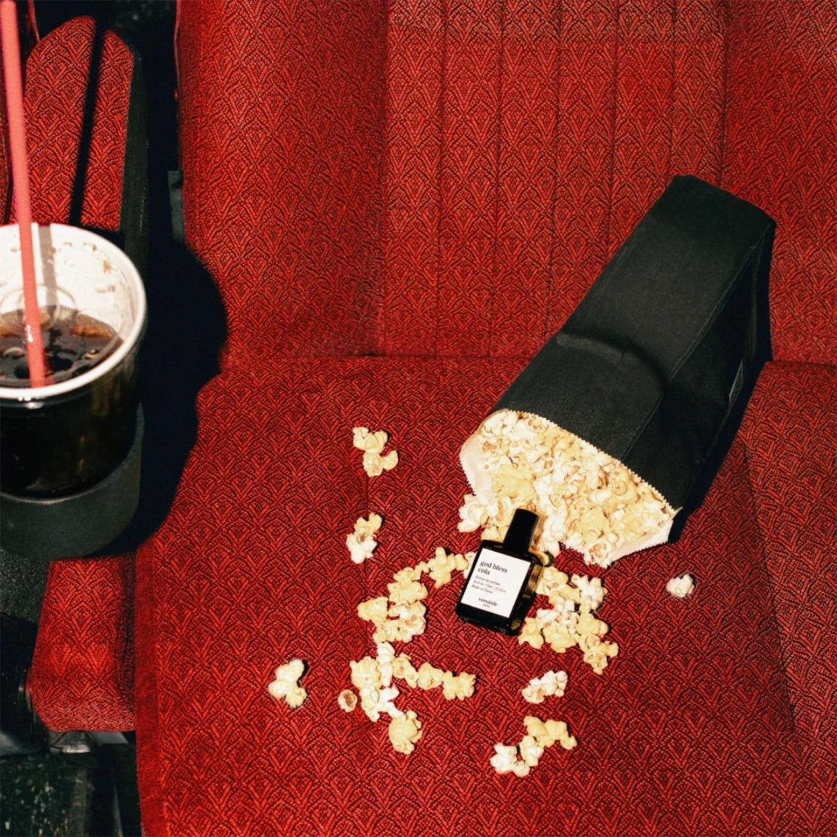 Versatile - God bless Cola Flakon umgeben von Popcorn auf rotem Kinosessel mit Cola in Becherhalterung