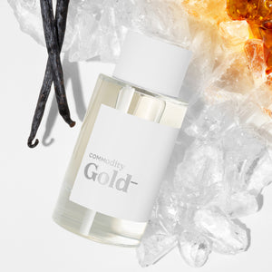 Commodity - Gold- Parfumflakon mit weißem Etikett umgeben von Vanilleschoten und Bernstein auf hellen Kristallen präsentiert