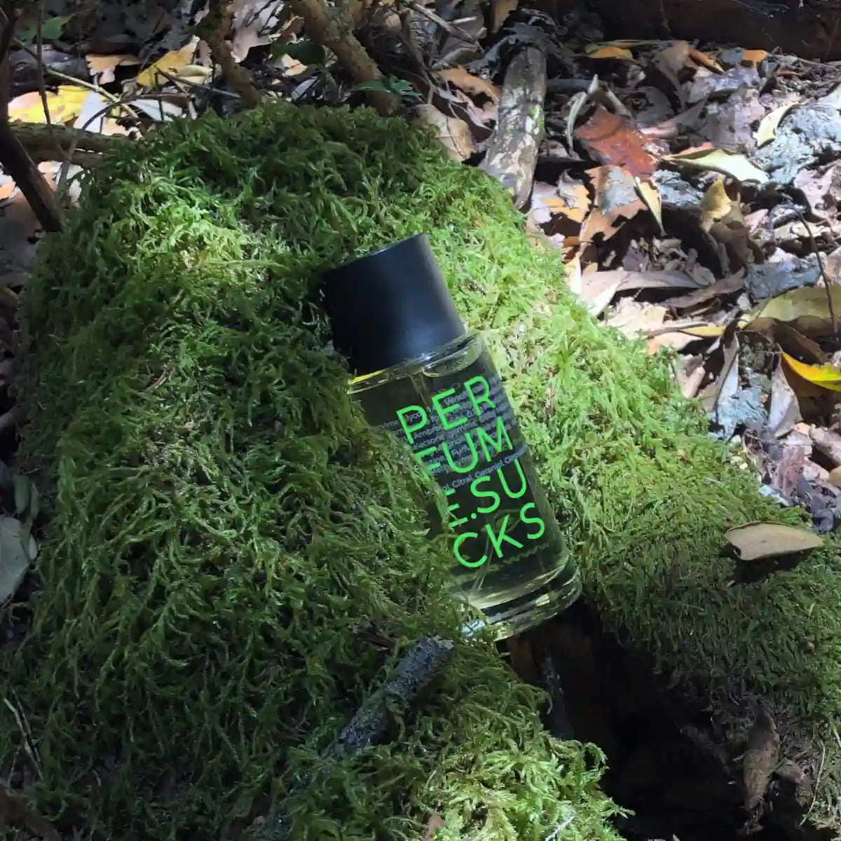PERFUME.SUCKS - Green 368C Parfumflakon mit schwarzem Deckel und neongrüner Schrift auf Moos im Waldlicht