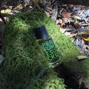 PERFUME.SUCKS - Green 368C Parfumflakon mit schwarzem Deckel und neongrüner Schrift auf Moos im Waldlicht
