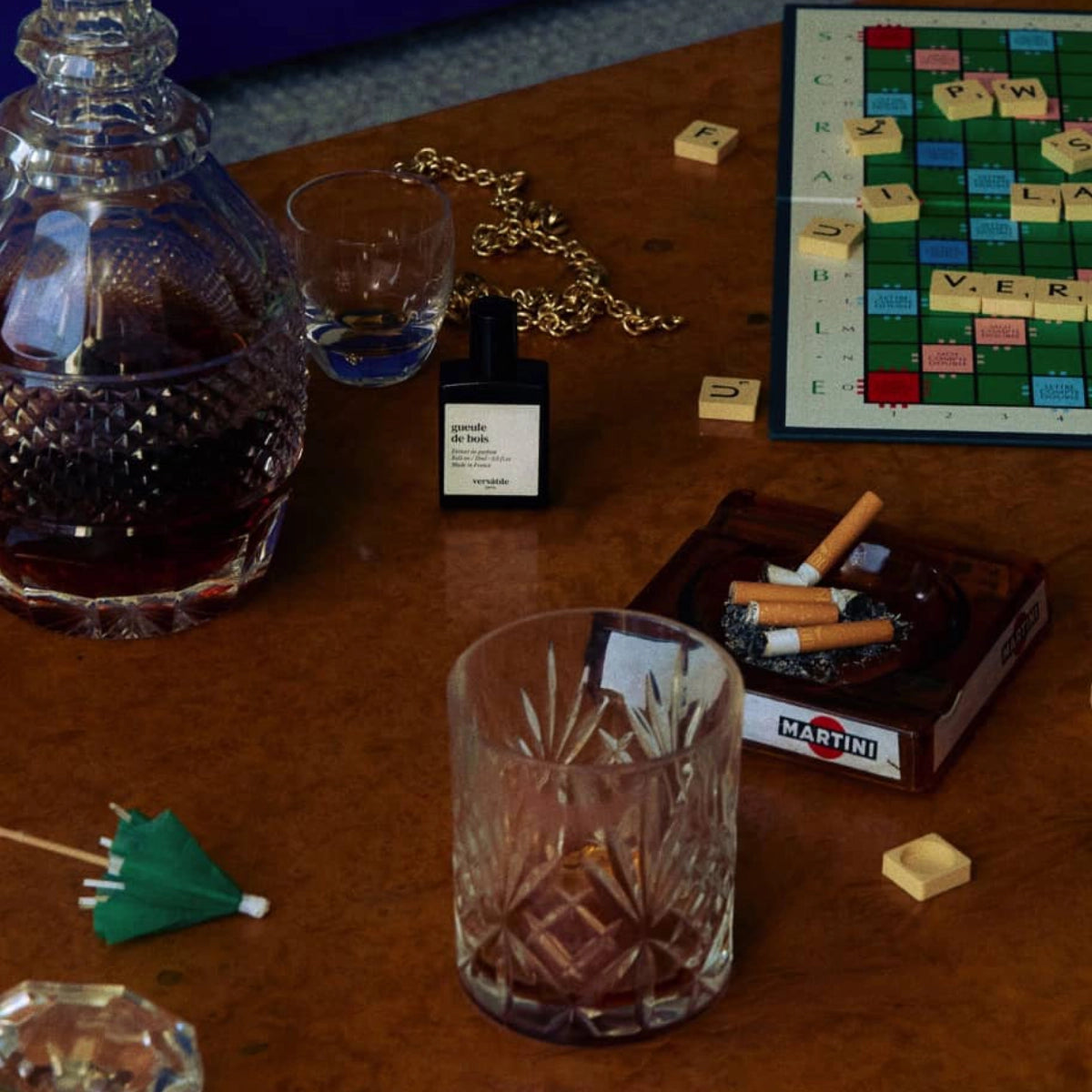 Versatile - Gueule de Bois Flakon neben Kristallglas, Kette, Scrabble-Steinen und Zigaretten im warmen Ambiente auf Holztisch