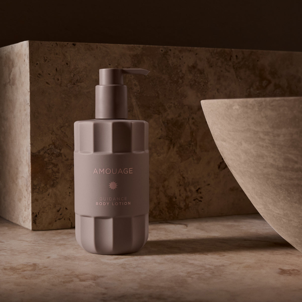 Amouage - Guidance - Body Lotion in modernem, mattgrauem Spender vor warmem Natursteinhintergrund